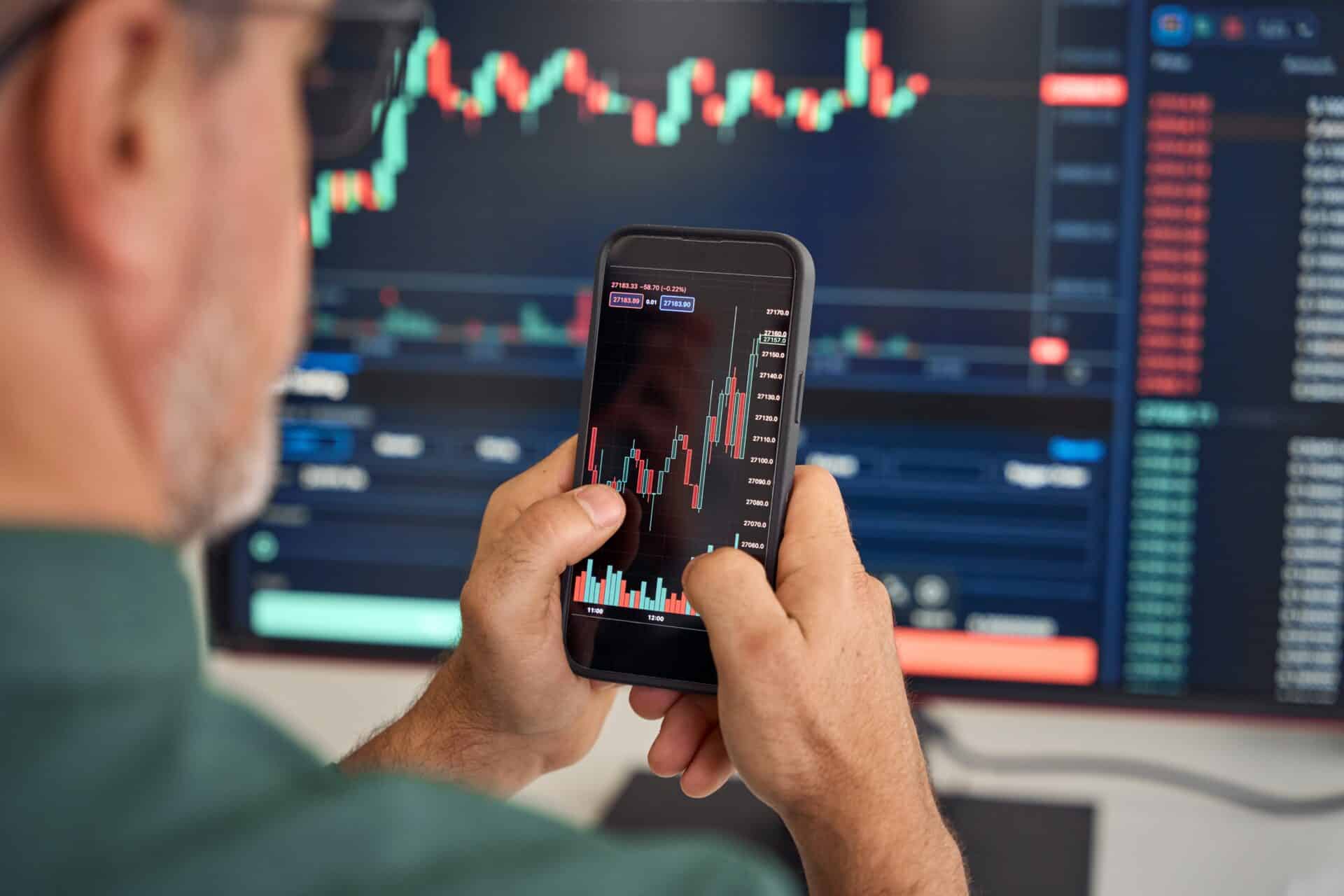 Investor analyzing stock market data on a smartphone, with financial charts displayed on a large screen in the background.